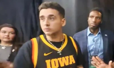 Young man wearing a black Iowa basketball jersey #22, looking to the left, in a backstage area with two people nearby.