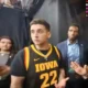 Young man wearing a black Iowa basketball jersey #22, looking to the left, in a backstage area with two people nearby.
