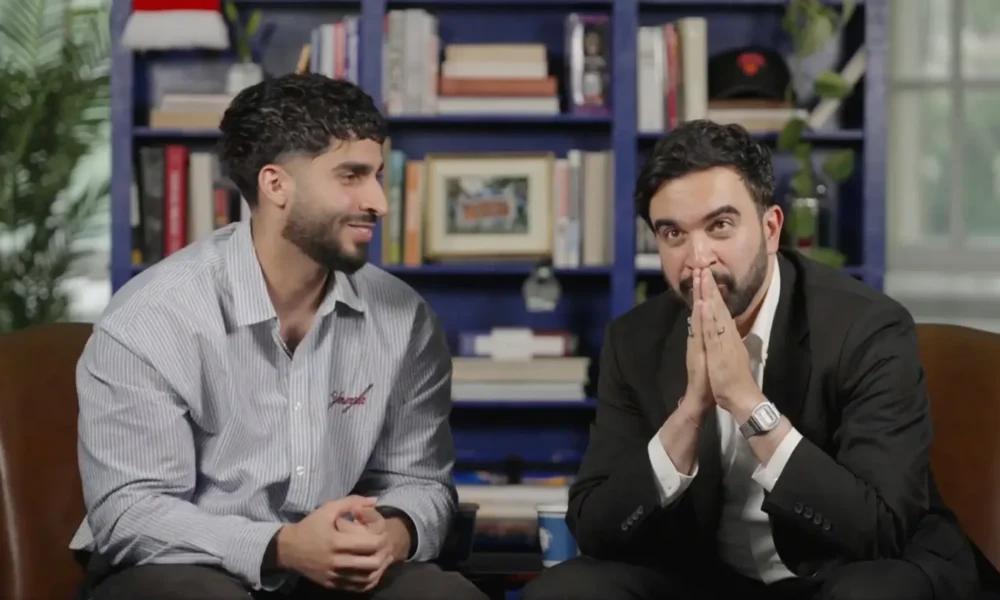 Two men sit on brown chairs in a book-filled room; one in a striped shirt smiles while the other in a black suit clasps his hands in front of his face as if praying or thinking.