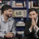 Two men sit on brown chairs in a book-filled room; one in a striped shirt smiles while the other in a black suit clasps his hands in front of his face as if praying or thinking.