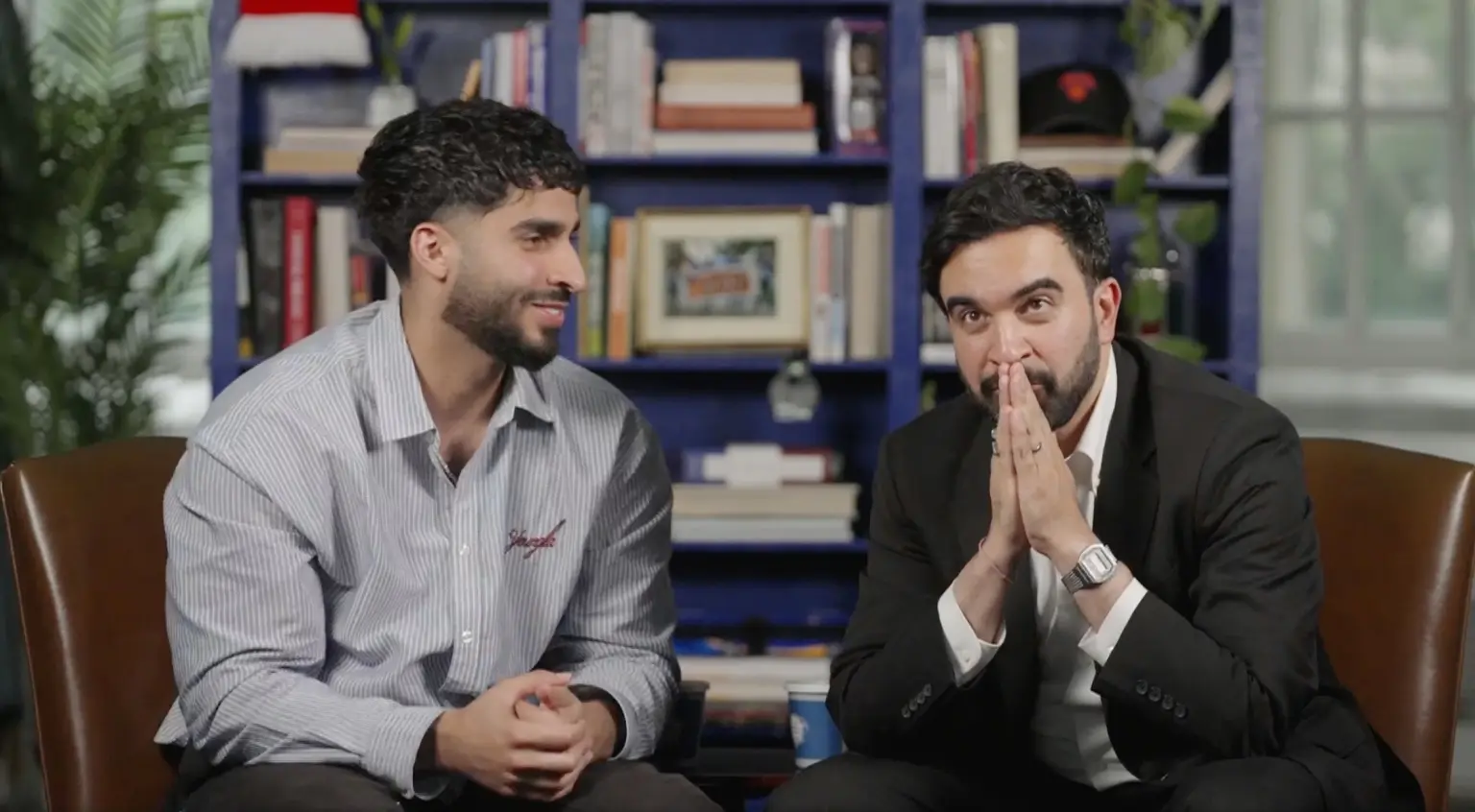 Two men sit on brown chairs in a book-filled room; one in a striped shirt smiles while the other in a black suit clasps his hands in front of his face as if praying or thinking.